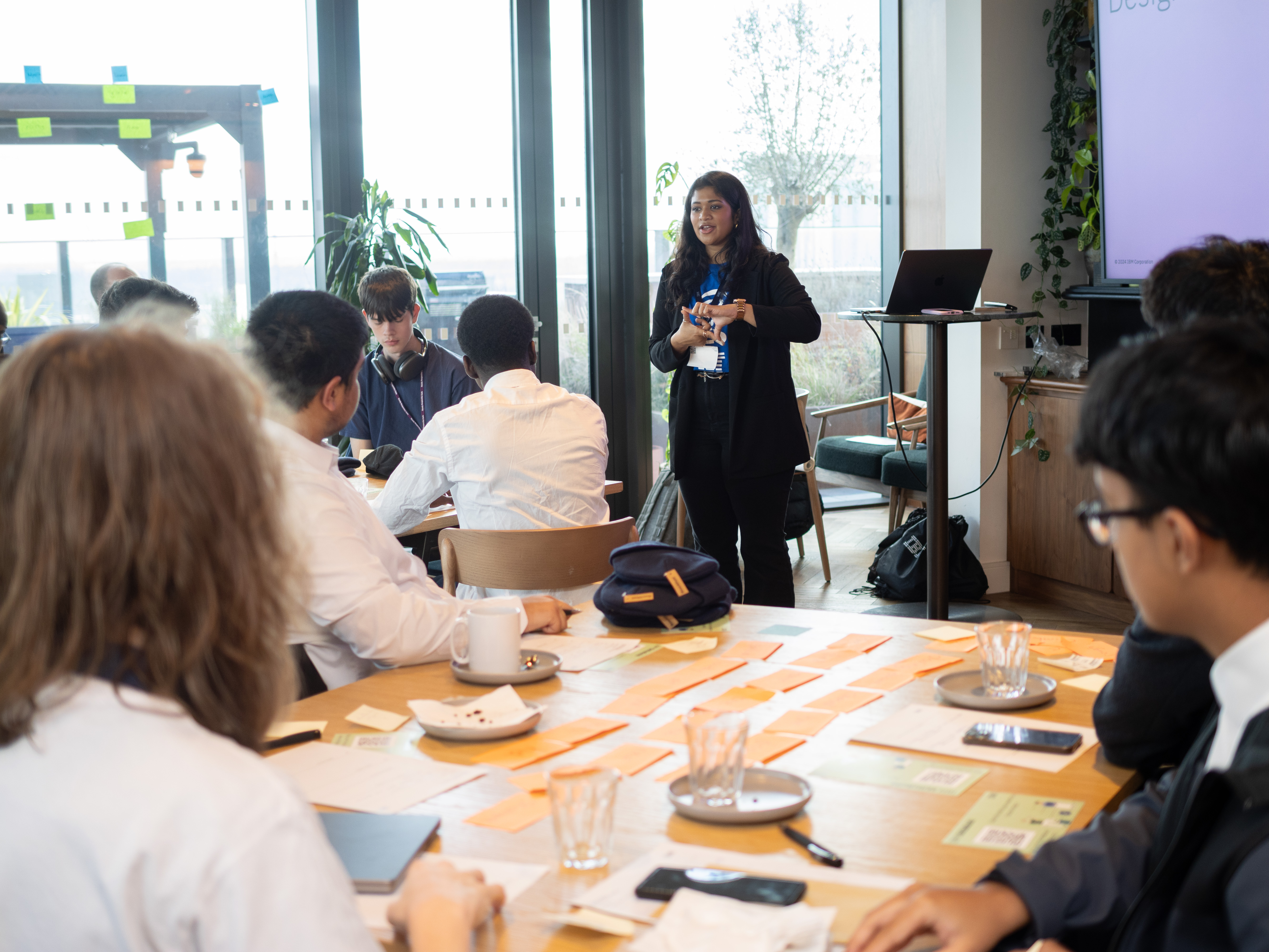 Karishma facilitating a design thinking workshop, presenting to participants seated around a table covered in activity cards