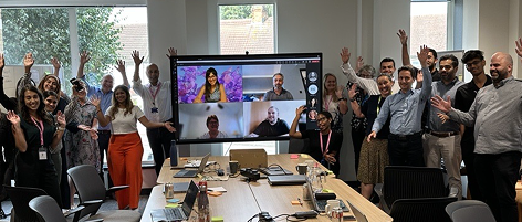 Workshop participants celebrating with raised hands around a table, joined by remote attendees on a video call screen